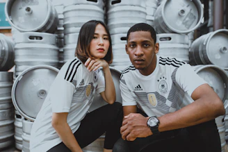 Two individuals wearing matching sports jerseys with a German logo are posing in front of several large metal barrels stacked up. They both have serious expressions and are sitting closely together.