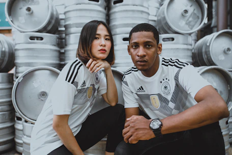 Two individuals wearing matching sports jerseys with a German logo are posing in front of several large metal barrels stacked up. They both have serious expressions and are sitting closely together.