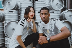 Two individuals wearing matching sports jerseys with a German logo are posing in front of several large metal barrels stacked up. They both have serious expressions and are sitting closely together.