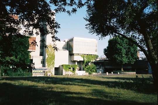 A modern building with large glass windows and geometric shapes is partially covered in green ivy. It sits in a landscaped area with grass and trees casting shadows in the foreground. The sky is clear and blue, contributing to a serene atmosphere.