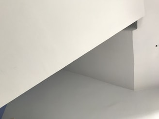 A minimalist architectural structure with intersecting angular lines and flat surfaces, primarily in shades of white and light gray. The image features a geometric composition with no visible furnishings or decorations.