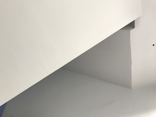 A minimalist architectural structure with intersecting angular lines and flat surfaces, primarily in shades of white and light gray. The image features a geometric composition with no visible furnishings or decorations.
