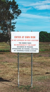 A warning sign in a rural area with text advising visitors to enter at their own risk, emphasizing not to approach or feed livestock and taking no responsibility for injuries. The landscape includes grassy fields and a tree under a blue sky with some clouds.