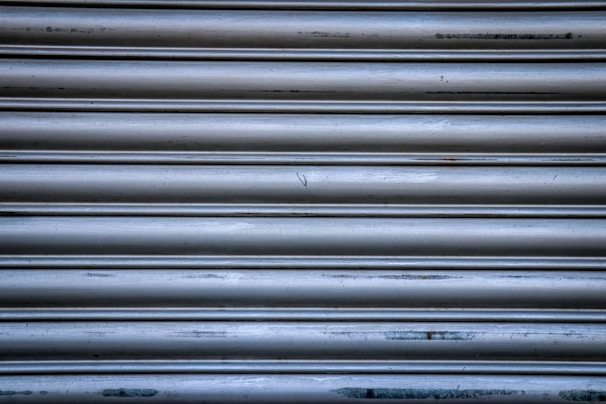 A close-up view of a metallic shutter with horizontal ridges and a slightly distressed appearance, featuring some scratches and discolorations on the surface.