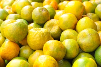 A large pile of fresh citrus fruits with smooth and textured skins, predominantly featuring shades of yellow and green. The fruits are tightly packed together, creating a vibrant and colorful display.