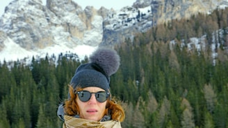 A rugged man wearing a thick, dark knitted hat standing against a snowy mountain backdrop.
