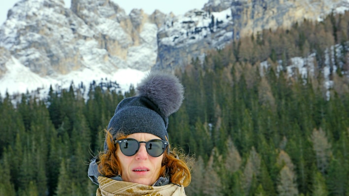 A rugged man wearing a thick, dark knitted hat standing against a snowy mountain backdrop.