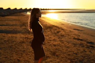 Pregnant woman standing on a rocky Galician coastline at sunset, soft light illuminating her silhouette.
