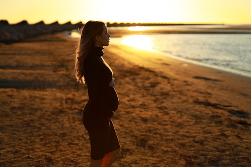 Pregnant woman standing on a rocky Galician coastline at sunset, soft light illuminating her silhouette.