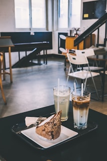 A cozy cafe setting with a slice of cake served on a white plate alongside two tall glasses filled with mixed beverages, topped with straws. The background features a minimalistic and modern interior with empty chairs, tables and large windows letting in natural light.