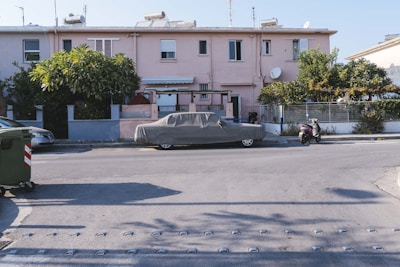 A residential street with a row of attached houses in the background, painted in light pink. The scene includes a covered car parked along the roadside, a motorcycle nearby, and a large green garbage bin on the left side. Two lush bushes flank the entrance to the house, which also has visible satellite dishes on the roof.