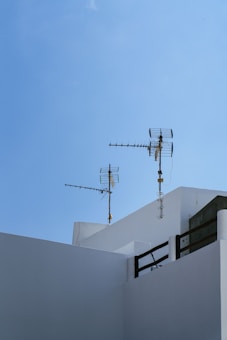 Antennas are mounted on the top of a minimalist white building against a clear blue sky. The architectural style is clean and geometric, with sharply defined edges.