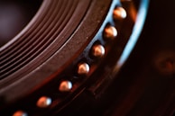 Close-up of a mechanical component with circular grooves and a row of metallic rivets. The image displays intricate details and textures, emphasizing the precision in engineering. Warm lighting highlights the metallic tones, adding depth and contrast to the scene.