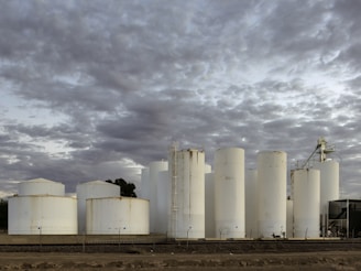 Several large, cylindrical white storage tanks stand against a backdrop of thick, overcast clouds. The tanks are arranged in a line with some metal ladders and pipes visible. The ground is barren and dark, indicating a possibly industrial setting.