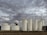 Several large, cylindrical white storage tanks stand against a backdrop of thick, overcast clouds. The tanks are arranged in a line with some metal ladders and pipes visible. The ground is barren and dark, indicating a possibly industrial setting.