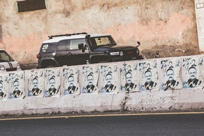 A row of campaign posters is prominently displayed on a concrete barrier, featuring a man's face with text in a non-English script. Behind the barrier, two cars are parked against a weathered wall with peeling paint.