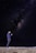 A person stands on a beach at night gazing at a smartphone, with the vast Milky Way galaxy stretching across the sky above. The starlit sky is filled with countless twinkling stars, providing a breathtaking cosmic backdrop.