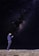 A person stands on a beach at night gazing at a smartphone, with the vast Milky Way galaxy stretching across the sky above. The starlit sky is filled with countless twinkling stars, providing a breathtaking cosmic backdrop.