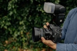 A person is holding a high-end DSLR camera equipped with a large lens and an external flash attachment. The camera is being stabilized with both hands, and the person is wearing a denim shirt. The surrounding background is lush greenery, slightly blurred to keep focus on the camera.