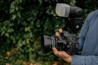 A person is holding a high-end DSLR camera equipped with a large lens and an external flash attachment. The camera is being stabilized with both hands, and the person is wearing a denim shirt. The surrounding background is lush greenery, slightly blurred to keep focus on the camera.
