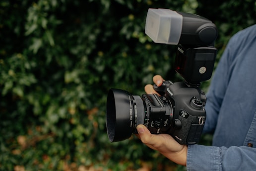 A person is holding a high-end DSLR camera equipped with a large lens and an external flash attachment. The camera is being stabilized with both hands, and the person is wearing a denim shirt. The surrounding background is lush greenery, slightly blurred to keep focus on the camera.