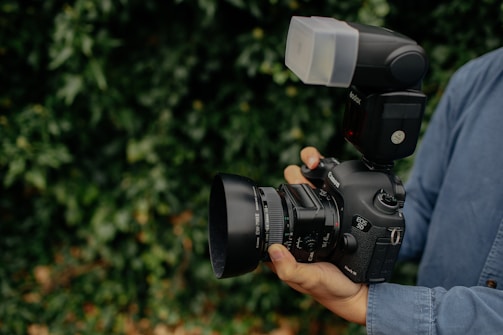 A person is holding a high-end DSLR camera equipped with a large lens and an external flash attachment. The camera is being stabilized with both hands, and the person is wearing a denim shirt. The surrounding background is lush greenery, slightly blurred to keep focus on the camera.