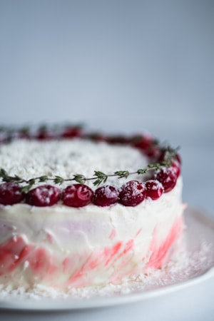 A beautifully decorated cake with a smooth white icing exterior, garnished with a border of bright red cherries and sprigs of green thyme. The lower part of the cake features pink marbled icing details, and there's a dusting of powdered sugar on top and around the cherries for a frosted look. The cake sits on a plain white plate and is captured against a neutral light gray background.
