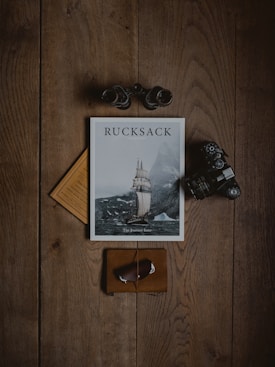 A wooden surface forms the background for a neatly arranged set of items. At the center is a magazine titled 'Rucksack', featuring an image of a ship amidst a cloudy and mountainous backdrop. Surrounding the magazine are binoculars, a vintage camera, a leather-bound notebook, and a yellow piece of paper.