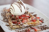 A decadent dessert featuring a golden waffle adorned with fresh strawberries and a generous scoop of vanilla ice cream. Chocolate sauce is drizzled over the top, adding richness to the presentation. The waffle is dusted with powdered sugar, enhancing its visual appeal.