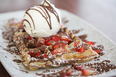 A decadent dessert featuring a golden waffle adorned with fresh strawberries and a generous scoop of vanilla ice cream. Chocolate sauce is drizzled over the top, adding richness to the presentation. The waffle is dusted with powdered sugar, enhancing its visual appeal.