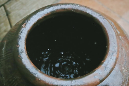 An overhead view of a clay pot filled with water, showing some ripples and droplets on the surface. The pot has a weathered look with a rough, textured surface.