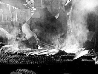 A person skillfully grills fish over an open flame, with smoke rising around, creating an atmospheric scene. The setting appears to be a food stall or restaurant kitchen.