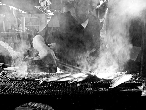A person skillfully grills fish over an open flame, with smoke rising around, creating an atmospheric scene. The setting appears to be a food stall or restaurant kitchen.