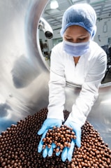 A worker in a laboratory coat, hairnet, and face mask is handling a large quantity of chocolate-coated products in a stainless steel drum using blue gloves. The setting appears to be a sterile environment, likely a food production or quality control facility.