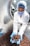 A worker in a laboratory coat, hairnet, and face mask is handling a large quantity of chocolate-coated products in a stainless steel drum using blue gloves. The setting appears to be a sterile environment, likely a food production or quality control facility.