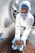 A worker in a laboratory coat, hairnet, and face mask is handling a large quantity of chocolate-coated products in a stainless steel drum using blue gloves. The setting appears to be a sterile environment, likely a food production or quality control facility.
