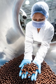 A worker in a laboratory coat, hairnet, and face mask is handling a large quantity of chocolate-coated products in a stainless steel drum using blue gloves. The setting appears to be a sterile environment, likely a food production or quality control facility.