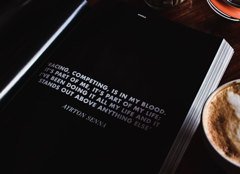 An open book lies on a wooden table next to a cup of cappuccino, showcasing a motivational quote by Ayrton Senna about racing and competing. The page is predominantly black with white text, and the quote is prominently displayed.