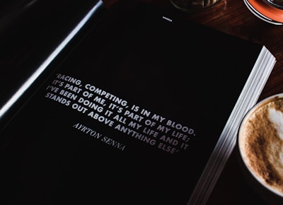 An open book lies on a wooden table next to a cup of cappuccino, showcasing a motivational quote by Ayrton Senna about racing and competing. The page is predominantly black with white text, and the quote is prominently displayed.