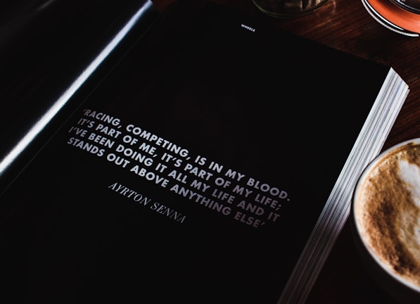 An open book lies on a wooden table next to a cup of cappuccino, showcasing a motivational quote by Ayrton Senna about racing and competing. The page is predominantly black with white text, and the quote is prominently displayed.