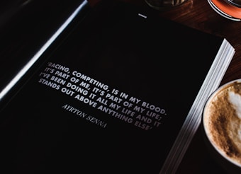 An open book lies on a wooden table next to a cup of cappuccino, showcasing a motivational quote by Ayrton Senna about racing and competing. The page is predominantly black with white text, and the quote is prominently displayed.