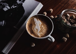 A cup of frothy coffee sits on a wooden table next to a jar filled with sugar cubes. Adjacent to it is an open book or magazine featuring a black and white photo of a person holding a cup. The lighting is dim and creates a cozy atmosphere.