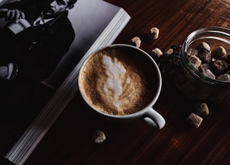 A cup of frothy coffee sits on a wooden table next to a jar filled with sugar cubes. Adjacent to it is an open book or magazine featuring a black and white photo of a person holding a cup. The lighting is dim and creates a cozy atmosphere.