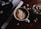 A cup of frothy coffee sits on a wooden table next to a jar filled with sugar cubes. Adjacent to it is an open book or magazine featuring a black and white photo of a person holding a cup. The lighting is dim and creates a cozy atmosphere.