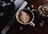 A cup of frothy coffee sits on a wooden table next to a jar filled with sugar cubes. Adjacent to it is an open book or magazine featuring a black and white photo of a person holding a cup. The lighting is dim and creates a cozy atmosphere.