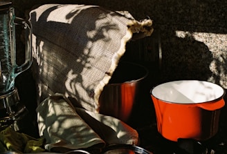 A kitchen scene with a blender, a red pot, and a metal bowl partially covered by a burlap cloth. There are shadow patterns cast on the burlap, creating an interplay of light and dark. A green cloth is casually draped over the counter.