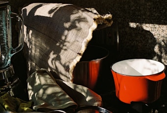 A kitchen scene with a blender, a red pot, and a metal bowl partially covered by a burlap cloth. There are shadow patterns cast on the burlap, creating an interplay of light and dark. A green cloth is casually draped over the counter.
