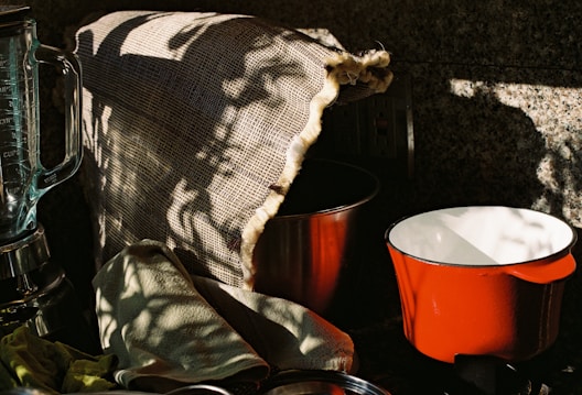A kitchen scene with a blender, a red pot, and a metal bowl partially covered by a burlap cloth. There are shadow patterns cast on the burlap, creating an interplay of light and dark. A green cloth is casually draped over the counter.