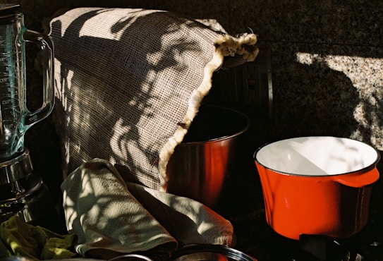 A kitchen scene with a blender, a red pot, and a metal bowl partially covered by a burlap cloth. There are shadow patterns cast on the burlap, creating an interplay of light and dark. A green cloth is casually draped over the counter.