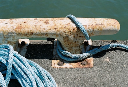 A rusty metal bollard is secured to a concrete surface with a blue rope wrapped around it. The water in the background is a deep greenish-blue, suggesting a dockside location.